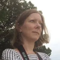 A person with shoulder length hair stands outside, wearing a striped shirt and holding a camera. Trees and a light sky are in the background.